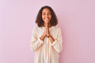 Young brazilian woman wearing striped shirt standing over isolated pink background praying with...