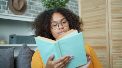 Pretty young African American woman is reading book at home turning pages smiling enjoying literature. Lifestyle, education and smart people concept.