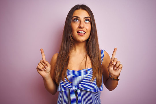Young beautiful woman wearing striped dress standing over isolated pink background amazed and surprised looking up and pointing with fingers and raised arms.