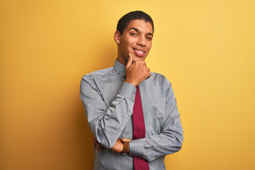 Young handsome arab businessman wearing shirt and tie over isolated yellow background looking confident at the camera smiling with crossed arms and hand raised on chin. Thinking positive.