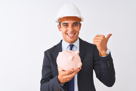 Handsome architect man wearing helmet holding piggy bank over isolated white background pointing and showing with thumb up to the side with happy face smiling