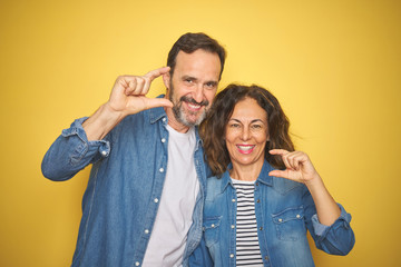 Fototapeta premium Beautiful middle age couple together wearing denim shirt over isolated yellow background smiling and confident gesturing with hand doing small size sign with fingers looking and the camera