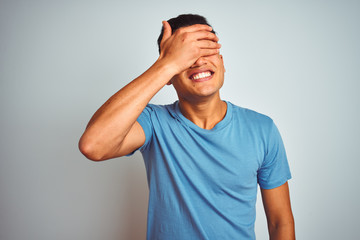 Fototapeta na wymiar Young brazilian man wearing blue t-shirt standing over isolated white background smiling and laughing with hand on face covering eyes for surprise. Blind concept.