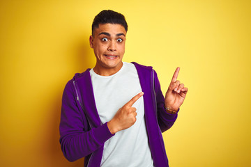 Young brazilian man wearing purple sweatshirt standing over isolated yellow background Pointing aside worried and nervous with both hands, concerned and surprised expression