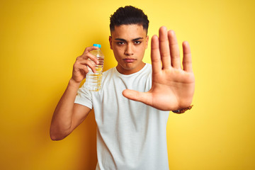 Young brazilian man holding bottle of water standing over isolated yellow background with open hand doing stop sign with serious and confident expression, defense gesture