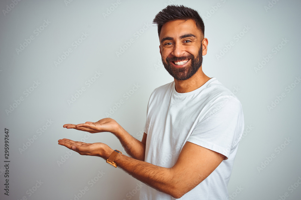 Wall mural Young indian man wearing t-shirt standing over isolated white background pointing aside with hands open palms showing copy space, presenting advertisement smiling excited happy