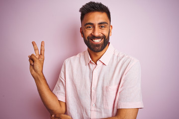 Fototapeta premium Young indian man wearing casual shirt standing over isolated pink background smiling with happy face winking at the camera doing victory sign. Number two.