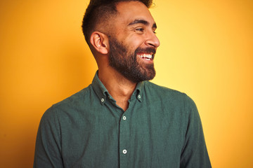 Fototapeta premium Young indian man wearing green shirt standing over isolated yellow background looking away to side with smile on face, natural expression. Laughing confident.