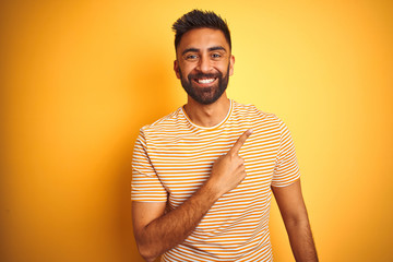 Young indian man wearing t-shirt standing over isolated yellow background cheerful with a smile on face pointing with hand and finger up to the side with happy and natural expression