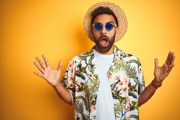 Indian man on vacation wearing floral shirt hat sunglasses over isolated yellow background...