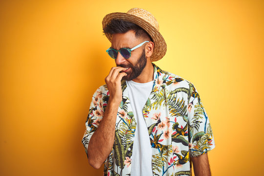 Indian man on vacation wearing floral shirt hat sunglasses over isolated yellow background looking stressed and nervous with hands on mouth biting nails. Anxiety problem.