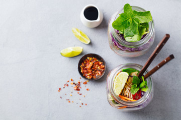 Noodles soba salad, soup with vegetables, tofu and chicken in jars. Grey background. Copy space. Top view.