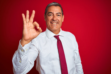 Handsome middle age businessman standing over isolated red background smiling positive doing ok sign with hand and fingers. Successful expression.