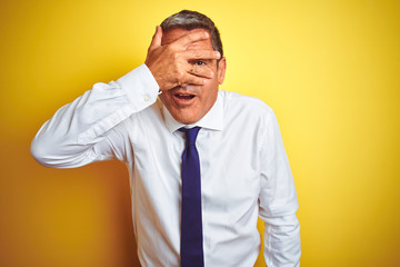 Handsome middle age businessman standing over isolated yellow background peeking in shock covering...