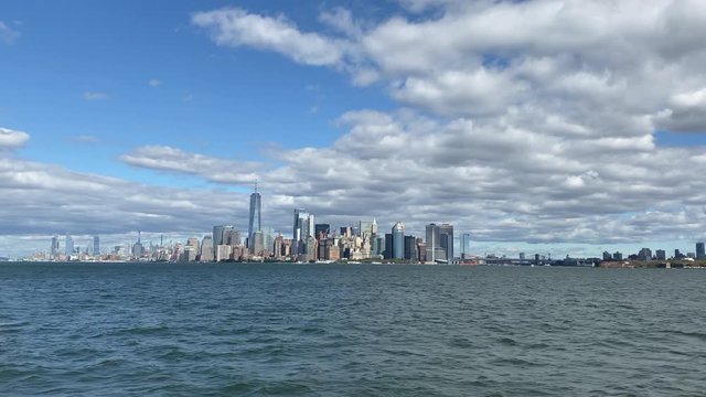 New York Manhattan skyline and waterfront filmed from cruise boat passing by. Wide establishing shot with blue sky and clouds.