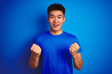 Fototapeta premium Young asian chinese man wearing t-shirt standing over isolated blue background celebrating surprised and amazed for success with arms raised and open eyes. Winner concept.