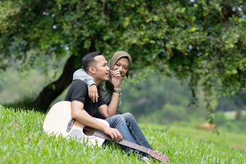 Asian muslim couple in love playing acoustic song guitar sitting on grass in the park under the tree