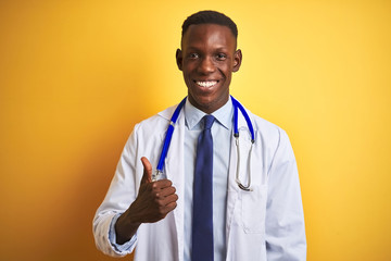 African american doctor man wearing stethoscope standing over isolated yellow background doing happy thumbs up gesture with hand. Approving expression looking at the camera with showing success.