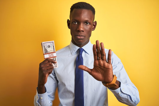 American businessman holding bunch of dollars standing over isolated yellow background with open hand doing stop sign with serious and confident expression, defense gesture
