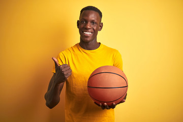 African american athlete man holding basketball ball standing over isolated yellow background happy with big smile doing ok sign, thumb up with fingers, excellent sign