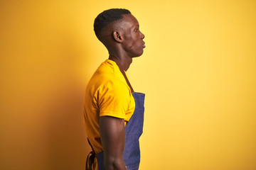 African american bartender man wearing apron standing over isolated yellow background looking to side, relax profile pose with natural face with confident smile.