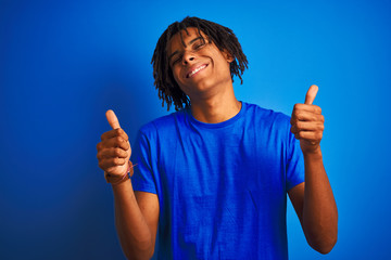 Afro american man with dreadlocks wearing t-shirt standing over isolated blue background success sign doing positive gesture with hand, thumbs up smiling and happy. Cheerful expression
