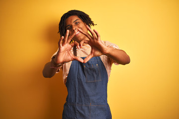 Afro american barista man with dreadlocks wearing apron over isolated yellow background smiling in love showing heart symbol and shape with hands. Romantic concept.
