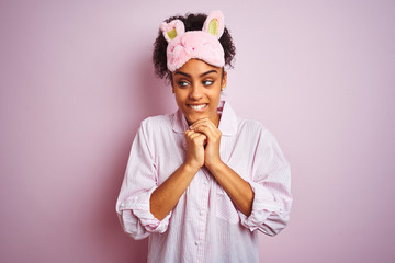 Young african american woman wearing pajama and mask over isolated pink background laughing nervous and excited with hands on chin looking to the side