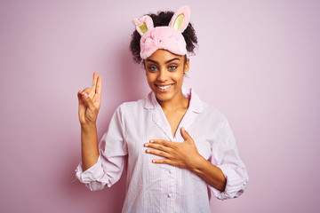 Young african american woman wearing pajama and mask over isolated pink background smiling swearing with hand on chest and fingers up, making a loyalty promise oath