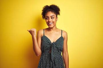 African american woman wearing summer casual green dress over isolated yellow background smiling with happy face looking and pointing to the side with thumb up.