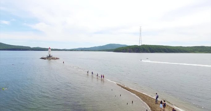 Flying above sand bar leading to the Tokarevsky lighthouse in Vladivostok. People sunbathe and walk on water to the lighthouse. Morot boat. Summer day. Russia. Aerial
