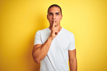 Young caucasian man wearing casual white t-shirt over yellow isolated background asking to be quiet with finger on lips. Silence and secret concept.