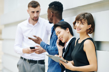 Pretty young smiling businesswoman standing outdoors with coworkers, holding tablet computer, leaning on wall and looking at camera