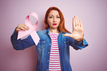 Young beautiful redhead woman holding cancer ribbon over pink isolated background with open hand...