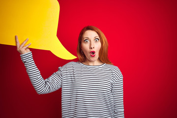 Young beautiful redhead woman holding yellow speech bubble over red isolated background scared in shock with a surprise face, afraid and excited with fear expression