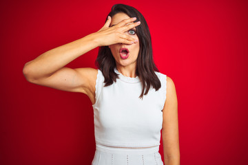 Young beautiful woman wearing white dress standing over red isolated background peeking in shock covering face and eyes with hand, looking through fingers with embarrassed expression.