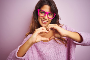 Young beautiful woman wearing fashion glasses standing over pink isolated background smiling in love showing heart symbol and shape with hands. Romantic concept.