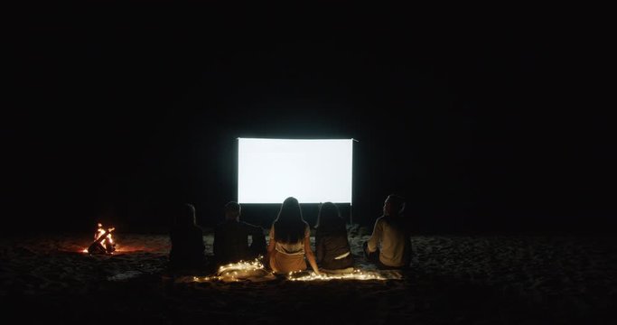 Open cinema. Friends gather around a campfire on the beach and watch a movie with a projector by the sea. White screen, place for your image and video. Open air cinema