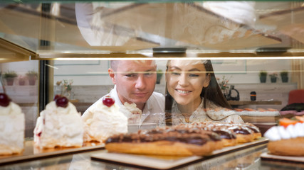 Fototapeta na wymiar happy young couple chooses tasty fresh cakes standing in glass show case of confectionery shop close view