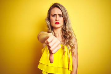 Young attactive woman wearing t-shirt standing over yellow isolated background looking unhappy and angry showing rejection and negative with thumbs down gesture. Bad expression.