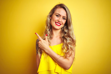 Young attactive woman wearing t-shirt standing over yellow isolated background cheerful with a smile of face pointing with hand and finger up to the side with happy and natural expression on face
