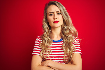 Young beautiful woman wearing stripes t-shirt standing over red isolated background skeptic and nervous, disapproving expression on face with crossed arms. Negative person.