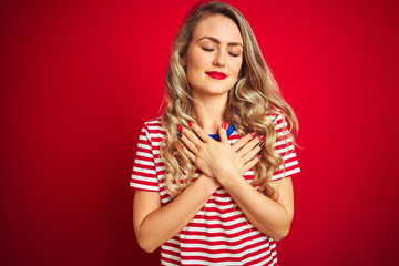 Young beautiful woman wearing stripes t-shirt standing over red isolated background smiling with hands on chest with closed eyes and grateful gesture on face. Health concept.
