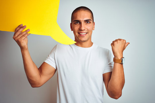 Young man holding yellow speech bubble over white isolated background pointing and showing with thumb up to the side with happy face smiling