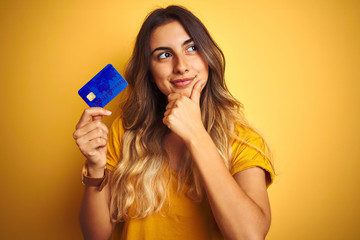Young beautiful woman holding credit card over yellow isolated background serious face thinking...
