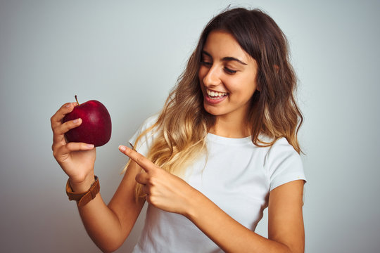 Young beautiful woman eating red apple over grey isolated background very happy pointing with hand and finger