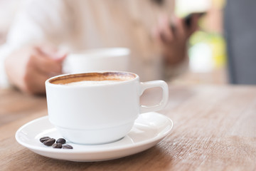 Hot coffee in the white cup and coffee beans in a sack on wooden table background with blurry woman holding coffee cup.Cafe drinking menu latte art coffee at the restaurant with copy space.