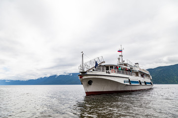 Large cruise ship during a walk on the water on the Teletskoye lake in the Altai mountains with tourists near the pier with rocks under the sky with clouds in the summer.