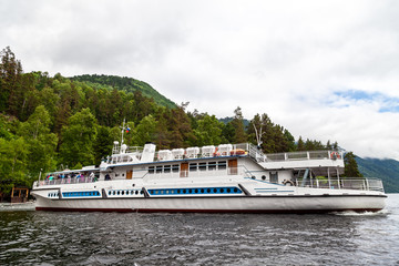 Obraz premium Large cruise ship during a walk on the water on the Teletskoye lake in the Altai mountains with tourists near the pier with rocks under the sky with clouds in the summer.
