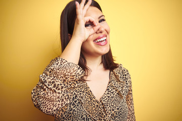 Young beautiful brunette woman wearing leopard shirt over yellow isolated background doing ok gesture with hand smiling, eye looking through fingers with happy face.